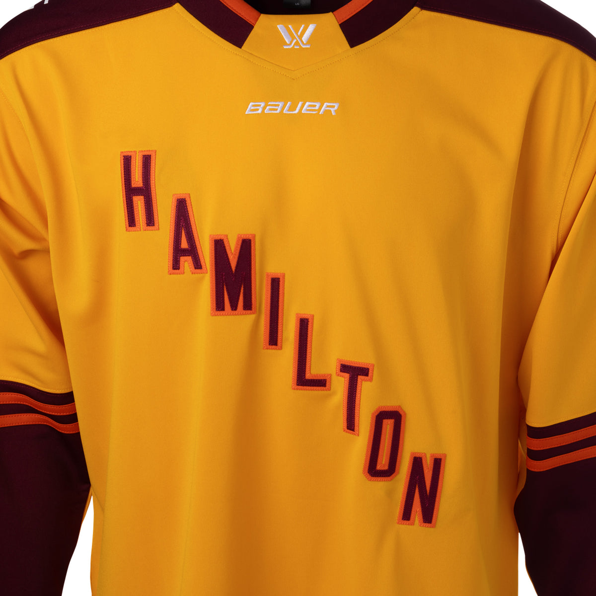 Front of a gold jersey with maroon shoulder caps. Maroon color blocking on the sleeves with orange stipes on top. 'HAMILTON' is in maroon with orange outlining diagonally across the chest. The collar is maroon with orange trim and a gold color block in front showcasing a white PWHL logo with a Bauer logo below it.