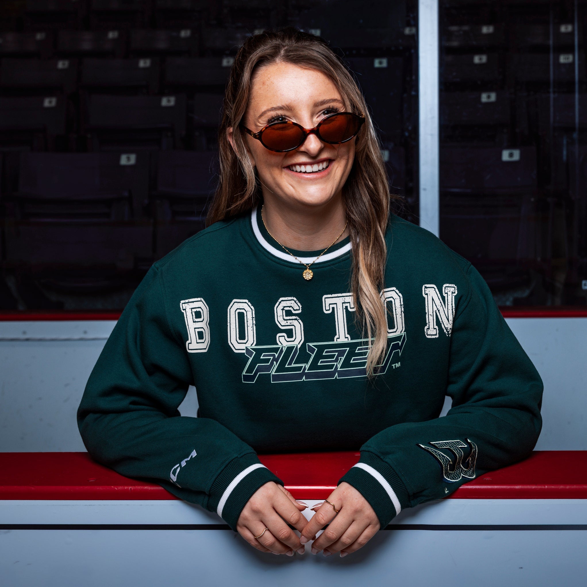Person wearing a green 'Boston Fleet' sweatshirt sitting on a bench in an indoor sports arena.