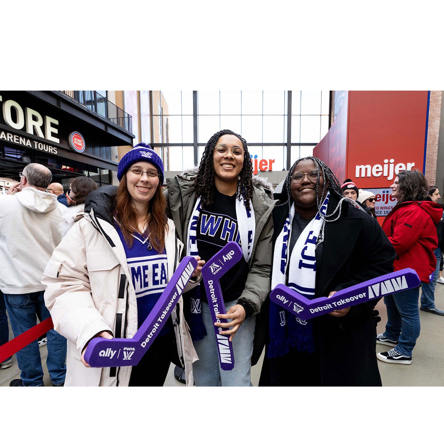Three women holding purple hockey sticks with sports team logos in a shopping mall.