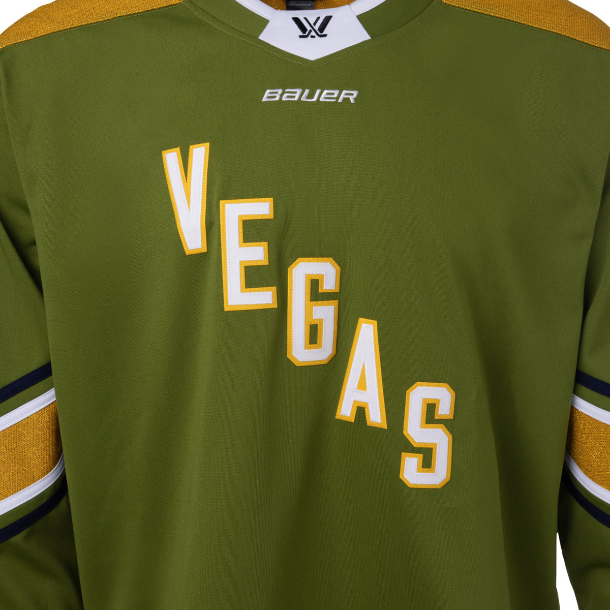 Front of a green jersey with sparkling gold shoulder caps and stripes along the sleeves surrounded by white and black stripes. In white outlined in gold reads 'VEGAS' diagonally across the chest. The collar is green with white trim on top. A black PWHL logo is centered on the collar with a white BAUER logo below it. 