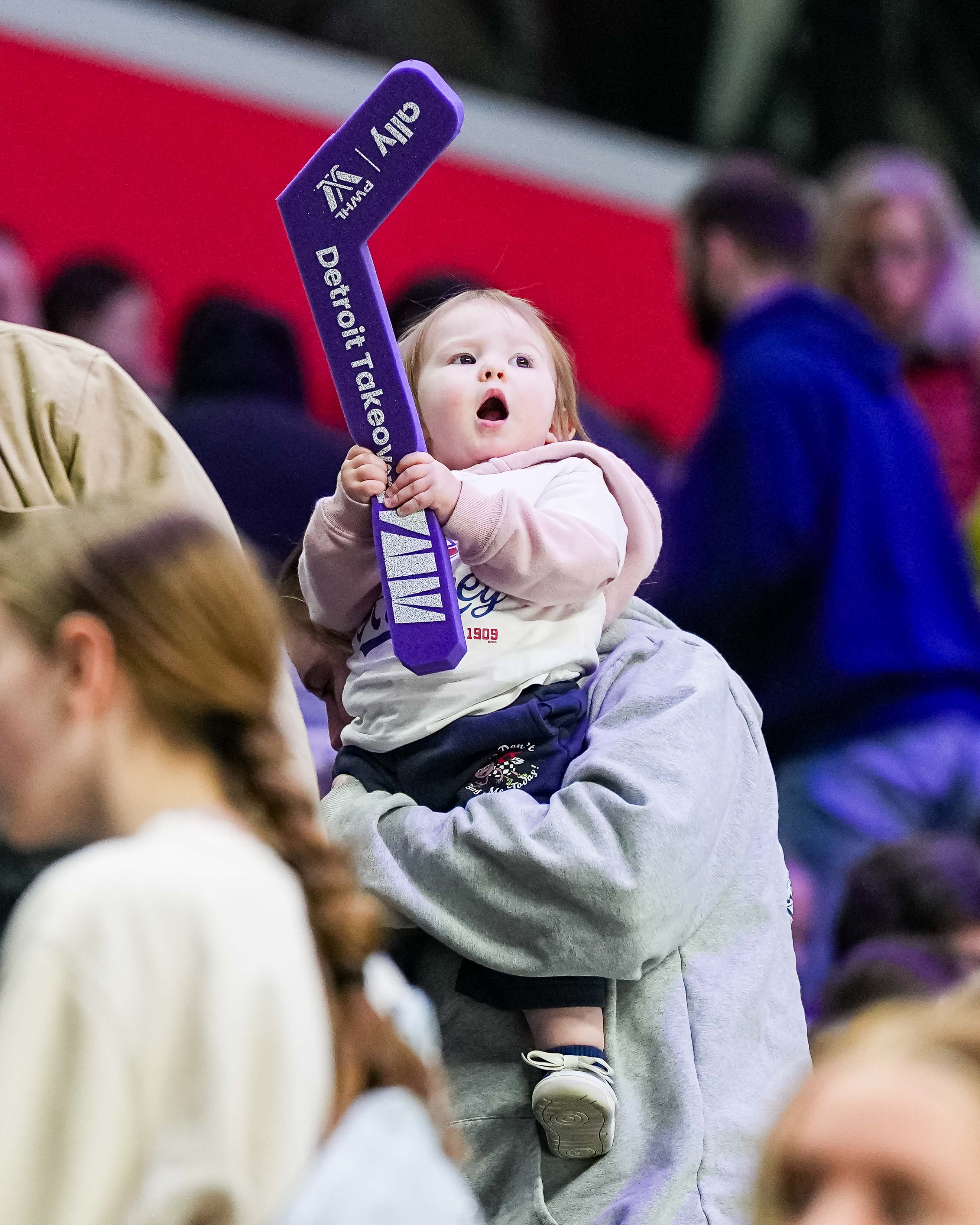 Child holding a purple hockey stick with 'Detroit Red Wings' branding in a crowd.
